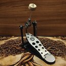 Černý bicí pedál s kovovou stoupací deskou v nerezovém provedení. Na desce je vyvrtáno několik otvorů a umístěno logo ve tvaru stylizovaného písmena…