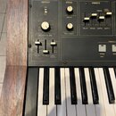 Detail ovládacího panelu a klávesnice elektronického nástroje v dřevěném rámu. Panel obsahuje knoflíky a posuvníky s nápisy OUTPUT, MASTER VOLUME, PI…