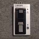 Stříbrný dvoupedálový spínací pedal BOSS DUAL FOOT SWITCH FS-7. Na pravé straně jsou písmena A a B s doprovodnými kontrolkami v červené a zelené barv…