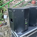 Dvě černé reproduktorové bedny s texturovaným povrchem umístěné na transportním kufru. Na horní hraně levého reproduktoru je nápis LOUDSPEAKER INSIDE…