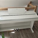 Bílé digitální piano s doprovodným stojanem a pedály. Na horním krytu je umístěno bílé zařízení s nápisem YAMAHA. Nástroj vykazuje čistý povrch bez v…