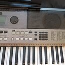 Stříbrný digitální klavír značky YAMAHA, model PSR E443. Nástroj má ovládací panel s displejem, množstvím tlačítek a seznamem stylů a hlasů. Povrch j…