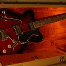 Polodupatá elektrická kytara s červeným sunburst povrchem a černým pickguardem leží v otevřeném kufru s oranžovou výplní. Nástroj má dva humbuckery,…