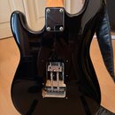 Zadní strana černé elektrické kytary s tvarem typu Stratocaster. V horní části je černý kryt elektroniky připevněný čtyřmi šrouby a ve spodní části j…