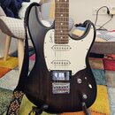 Elektrická kytara s tmavě hnědým träsováním těla a bílým pickguardem. Na pickguardu je viditelné logo Godin. Kytara má tři snímače, chromovaný most a…