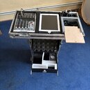 Otevřený transportní kufr s vnitřním pěnovým polstrováním, který obsahuje malý mixážní pult a bílý tablet. Na mixéru jsou viditelné posuvníky, knoflí…