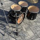 Černá bubnová sada s dřevěnými vnitřky korpusů, která obsahuje basový buben, dva tom-tomy a jeden floor tom. Na čínelu i na přední коже basového bubn…