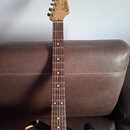 Elektrická kytara s hnědým gradientem těla a bílým pickguardem. Na hlavě z přírodního dřeva je viditelné logo Squier. Hmatník je tmavého dřeva s bílý…