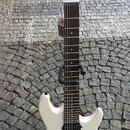 Bílá elektrická kytara s hnědým hmatníkem, umístěná na černém stojanu. Na hlavě je vidět nápis Sterling a Cutlass. Kytara má tři černé snímače, černý…