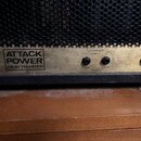 Detail spodní části kytarového zesilovače se zlatým kovovým panelem a černou mřížkou. Na panelu je nápis ATTACK POWER HEAVYMASTER a dva výstupy označ…