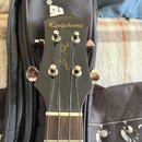 Černá hlava basové kytary s nápisem Epiphone a zlatým nápisem Les Paul MODEL. Nástroj má čtyři chromované ladičky a je umístěn před černým pouzdrem.…