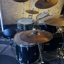 Celá bicí souprava v černé barvě s kovovým hardwarem. Na činelech jsou viditelné nápisy Zildjian a SABIAN (model HH X-Pliance). Souprava obsahuje bas…