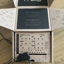 Kartonová krabice značky moog, uvnitř které se nachází syntezátor s bílým předním panelem. Na panelu jsou viditelné nápisy PRESET: SMOOTH BASS, VCO,…