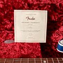 Bílý papírový certifikát s nápisem „GENUINE PRODUCT OF FENDER“, logem „Fender“ a textem „CERTIFICATE of AUTHENTICITY“. Jsou na něm uvedena čísla 0110…