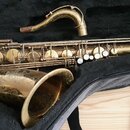 Zlatý saxofon v černém pouzdře, vedle kterého leží samostatný hnítek. Nástroj vykazuje viditelné opotřebení povrchového lakování a přítomnost škrában…