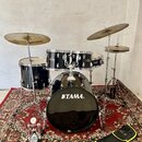 Černá bicí souprava s kovovým kováním, umístěná na červeném orientálním koberci. Na basovém bubnu je bílý nápis TAMA®. Souprava obsahuje tři tommy, s…