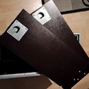 Dvě tmavě hnědé, pravděpodobně dřevěné desky s kovovými úchytkami v horní části. Desky jsou umístěny na kovovém rámu, který leží na světlé podlaze. N…