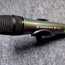 Černý bezdrátový mikrofon s kovovou mřížkou. Na těle je bílý nápis SENNHEISER a označení EW 100 G2. V dolní části je viditelná zelená nálepka s nečit…