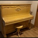 Světle hnědé dřevěné piano s ozdobným vyřezáváním v horní části. Před nástrojem stojí jednoduchá dřevěná taburetka na čtyřnohém podstavci. Povrch pia…