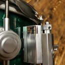 Detail kovového mechanismu na zeleném bubnu. Na kovovém krytu je bílá nálepka s nápisy „dw“, „Delta Throw Off“ a drobným textem začínajícím „U.S. Pat…