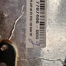 Detail kovového povrchu s nalepenou bílou štítkovou nálepkou. Na nálepce je čárcový kód a texty 7707406 - 000 oraz PG ASSY AM STD STRAT HSS SHAW PBP.…