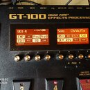 Černý efektový procesor s nápisem GT-100 COSM AMP EFFECTS PROCESSOR. Zařízení má dvě oranžové displeje s texty U01-4, Solo a dalšími zkratkami efektů…