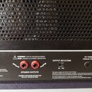 Zadní strana černého zesilovače s perforovaným kovovým krytem v horní části. Jsou zde dva červené konektory s nápisem SPEAKER OUTPUTS a přepínač OUTP…