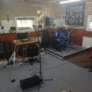 Fotografie zachycuje interiér hudebního studia s různým vybavením. V prostoru je vidět modrá bicí souprava, elektrická kytara s bílým tělem a červená…