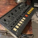 Černý kytarový syntetizátor s nápisem KORG GUITAR SYNTHESIZER. Zařízení má numerous otočné ovladače, přepínače a řadu žlutých a bílých tlačítek. Na p…