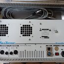 Bílý zesilovač PowerPack10 značky SkyTronic umístěný v hliníkovém kufru spolu s šedým kabelem. Na předním panelu jsou dva výstupy LEFT OUTPUT a RIGHT…