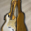Bílá elektrická kytara s tvarem Stratocaster v černém kufru s žlutým vnitřkem. Na hlavě hředu je viditelné logo Fender. Kytara má tři snímače, zlatý…