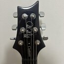 Černá hlava elektrické kytary s chromovanými ladičkami. Na povrchu je bílý nápis „Paul Reed Smith SE“ a slovo „Custom“. Povrch je hladký a lesklý.