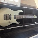 Světle šedá elektrická kytara v černém tvrdém kufru. Na hmatníku je viditelné logo LX. Kytara má dva humbuckery, tremolo most a jeden ovládací knoflí…
