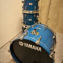 Sada modrých bubnů s kovovým kováním. Na předním černém obalu basového bubnu je bílý nápis YAMAHA s doprovodným logem. Na dvou stojících bubnech jsou…