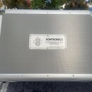 Stříbrný transportní kufr s texturovaným povrchem a kovovými hranami. Uprostřed je umístěna bílá nálepka s nápisem SONTRONICS PROFESSIONAL AUDIO EQUI…