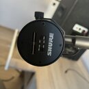 Černý kulatý mikrofon s nápisem SHURE. Na přední straně jsou vidět dvě úzké svislé rýhy a čtyři malé vkovy. Mikrofon je připevněn k šedému stativu po…