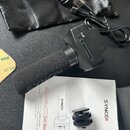 Černý bezdrátový mikrofon s pěnovou čepičkou na hlavě a ovládacím prvkem v horní části. Vedle mikrofonu leží uživatelský manuál s nápisem SYNCO))) a…