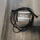 Kovová stůpková jednotka s dvěma spínacími tlačítky a připojeným černým kabelem. Na horní straně je logo Fender a nápisy CH SELECT a EFFECTS pod tlač…