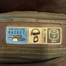Detail štítku na vnitřní straně transportního obalu z tmavého textilu. Na štítku jsou nápisy PROTECTION RACKET, CORNWALL • UK a rok 2014. Je zde také…