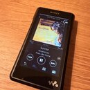 Černý digitální přehrávač značky SONY s displejem. Na obrazovce je zobrazen album cover Norah Jones, název skladby Sunrise a album Feels Like Home. V…