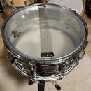Krytý snare drum s průhlednou blánou značky REMO, na které je nápis REMO® WEATHERKING MADE IN U.S.A. Na bláně je umístěn kovový pružinový mechanismus…