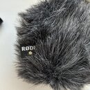 Šedý chlupatý kryt na mikrofon (tzv. deadcat). Na černé tkanině je vyšívaný bílý nápis RØDE. Povrch je rovnoměrně zastřižený, bez viditelných poškoze…