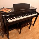 Černý digitální piano v podobě konzole s doprovodnou lavicí. Na přední straně nad klávesami je bílý nápis YAMAHA a vlevo u kláves menší nápis Clavino…