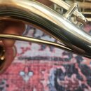 Detailní záběr na hradek a horní část těla saxofonu v lesklém zlatavém povrchu. Na pravé straně je vidět korkový spojovací prvek. Povrch nástroje vyk…