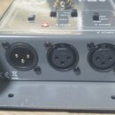 Šedý kovový přístroj s panelem ovládání a třemi XLR konektory na zadní straně. Konektory jsou označeny nápisy OUTPUT, INPUT B a INPUT A. Na horním pa…