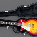 Elektrická kytara s červeným gradientem (sunburst) v černém tvrdém kufru. Na hlavě kytary je viditelné logo Vintage. Nástroj má dva humbuckery, bílý…
