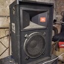 Černý reproduktor v bedně potažené tmavým kobercem s kovovými hranami. V horní části je rohové hrdlo s oranžovým logem JBL, ve spodní části je kruhov…