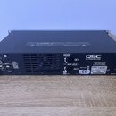 Šedý rackový zesilovač s nápisem QSC PLX POWER AMPLIFIER. Na zadní straně jsou vstupy označené jako INPUTS (CH 1, CH 2) a výstupy OUTPUT (CH 1, CH 2)…