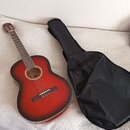 Akustická kytara s červeným gradientním povrchem korpusu a hnědým hřidnicem. Kytara je umístěna vedle černého měkkého pouzdra na kytaru. Na nástroji…