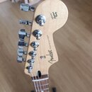 Záběr na hlavu elektrické kytary z přírodního dřeva s chromovanými ladičkami. Na zadní straně jsou nápisy Fender, STRATOCASTER® a ORIGINAL Contour Bo…